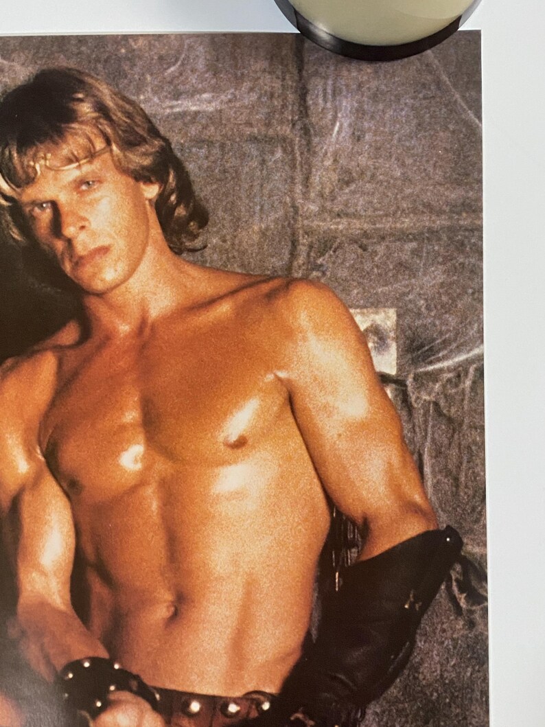 May include: A shirtless man with long blond hair poses for a photo. He is wearing a leather belt with studs and a leather wrist cuff. The background is a stone wall.