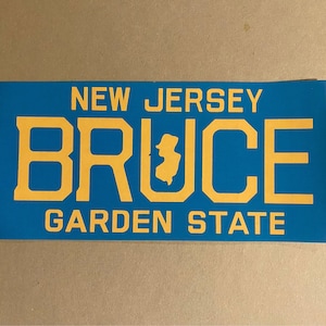 Puede incluir: Un letrero azul y amarillo con el texto "NEW JERSEY BRUCE GARDEN STATE" y un contorno del estado de Nueva Jersey.