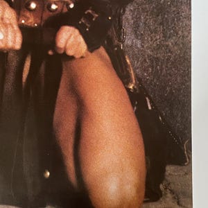 May include: A close-up of a person wearing a brown leather skirt and a black leather jacket. The person is kneeling on a sandy surface.