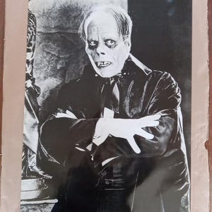 May include: Black and white photograph of a man in a suit and a mask. The man has a pale face and dark eyes. He is looking directly at the camera. The photograph is from the 1920s.