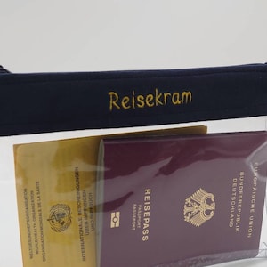 Könnte beinhalten: Eine transparente Reise-Tasche aus Plastik mit einem dunkelblauen Reißverschluss oben. Die Tasche ist mit "Reisekram" in goldener Stickerei beschriftet. Die Tasche enthält einen gelben und weißen internationalen Impfpass und einen burgunderfarbenen deutschen Reisepass.