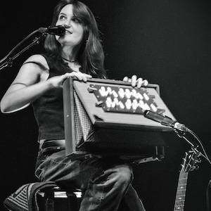 May include: A black and white image of a musician performing on stage. The musician is seated and holding a small, rectangular instrument with multiple white keys. A microphone is positioned in front of the musician, and a guitar is visible to the side.