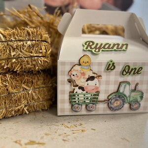 May include: A white favor box with a gingham pattern and farm animal illustrations. The box has a handle and says "Ryann is One" on the front.