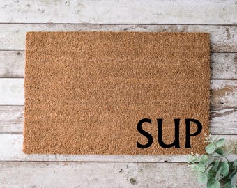 May include: A brown coir doormat with the word "SUP" printed in black letters.