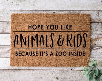 May include: A rectangular doormat made of natural coir fiber. The mat features the text "HOPE YOU LIKE ANIMALS & KIDS BECAUSE IT'S A ZOO INSIDE" in black, centered on the mat. The background is a light brown color.