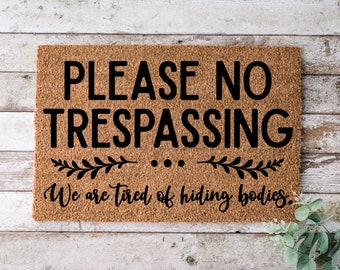 Könnte beinhalten: Eine braune Kokosmatte mit dem Text "PLEASE NO TRESPASSING... We are tired of hiding bodies." in schwarzer Schrift.