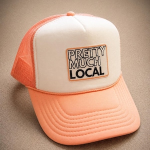May include: A peach-colored trucker hat featuring a white foam front panel with a rectangular patch that reads PRETTY MUCH LOCAL in black lettering. The cap includes a matching peach mesh back, a curved brim, and a decorative cord across the base.