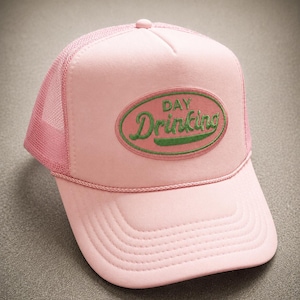 May include: A light pink trucker hat featuring a mesh back and a solid front panel. An oval patch is centered on the front, displaying the green embroidered text DAY Drinking. A matching pink cord trim runs horizontally across the base of the crown.