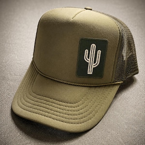 May include: An olive green trucker hat featuring a structured front panel with a rectangular patch displaying a white embroidered cactus graphic. The cap includes a matching olive green mesh back, a curved brim with decorative stitching, and a thin cord.