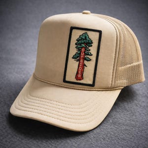 May include: Tan trucker hat with a mesh back and a curved brim. The front panel features a rectangular patch with a tree design and the word "OUTSIDER" in red. The hat has a beige rope detail and a button on top.