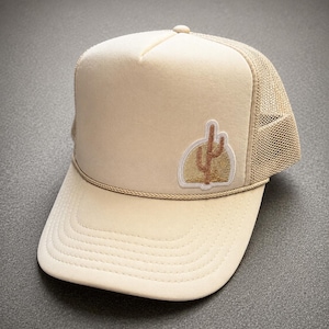 May include: A beige trucker hat featuring a mesh back, a structured front panel, and a decorative cord across the brim. An embroidered patch on the front displays a brown cactus silhouette set against a semi-circular gold background.