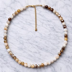 Può includere: Collana di perline con perline rotonde in varie tonalità di terra, tra cui bianco, marrone e beige. La collana ha una catena e una chiusura color oro. Le perline sembrano essere di pietra o materiale simile.