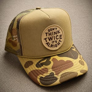 May include: A tan trucker hat featuring a mesh back, a camouflage print brim, and a circular patch on the front panel. The patch is tan with dark brown embroidered text that reads, "DON'T THINK TWICE IT'S ALRIGHT," and a yellow cord trim.