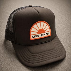 May include: A black trucker hat featuring a mesh back and a solid front panel. An embroidered patch on the front displays a stylized orange and white sun graphic above the text "LIVE SIMPLY" in black capital letters on a white background.