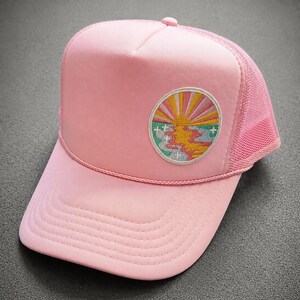 May include: A light pink trucker hat featuring a foam front panel, a mesh back, and a decorative cord across the brim. An embroidered circular patch on the front displays a stylized sunburst design with yellow, orange, and pink rays over a landscape.