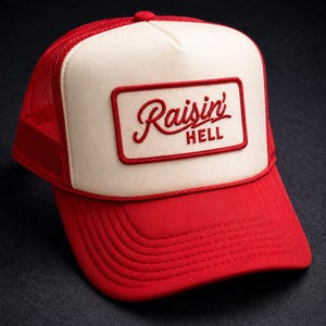 May include: A red and cream trucker hat featuring a rectangular patch on the front panel with the embroidered red text Raisin' HELL. The cap has a solid red brim, a cream-colored front panel, and a red mesh back with a matching red button on top.