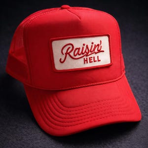 May include: A red trucker hat with a white rectangular patch. The patch features the words "Raisin' Hell" in a cursive font. The hat has a mesh back and a curved brim.