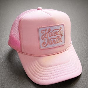 May include: A light pink trucker hat featuring a rectangular patch on the front panel. The patch has a light purple background with the text Howdy Darlin embroidered in a cursive pink font. The hat includes a mesh back and a matching pink brim.