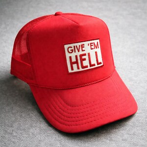 May include: A red trucker cap with a mesh back and a curved peak. The front of the cap has a white rectangular patch with the words "GIVE 'EM HELL" in white capital letters.
