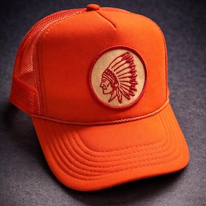 May include: Bright orange trucker hat with a mesh back and a curved brim. The hat features a circular patch with an embroidered Native American headdress design in red and tan. A stylish accessory for outdoor activities.
