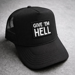 May include: A black trucker hat featuring a rectangular patch on the front panel with the white embroidered text GIVE 'EM HELL. The cap has a solid front, a mesh back, a curved brim, and a decorative cord detail across the base of the crown.