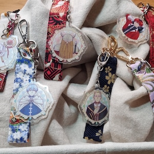 Può includere: Una collezione di portachiavi con illustrazioni di personaggi. Ogni portachiavi presenta un diverso design di personaggio, attaccato a cinturini in tessuto fantasia con chiusure in metallo. I cinturini in tessuto hanno disegni floreali, geometrici e fantasia. I portachiavi sono esposti in un cestino.