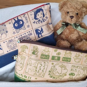 May include: Three fabric zippered pouches with Japanese characters and illustrations. The pouches are beige with blue trim, beige with green trim, and beige with brown trim. A brown teddy bear is sitting on top of the pouches.
