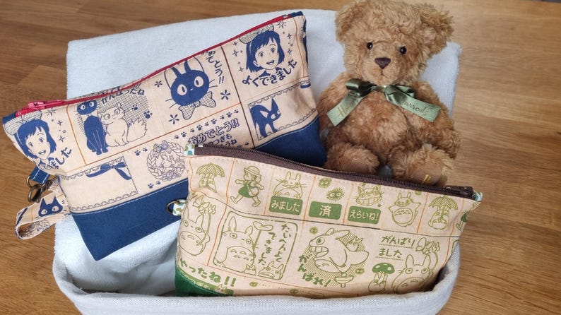 May include: Three fabric zippered pouches with Japanese characters and illustrations. The pouches are beige with blue trim, beige with green trim, and beige with brown trim. A brown teddy bear is sitting on top of the pouches.