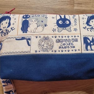 May include: A blue and cream fabric pouch with a zipper closure. The pouch features a repeating pattern of black cats, a white cat, and Japanese text. The text reads "できた!" and "おめでとう!!".