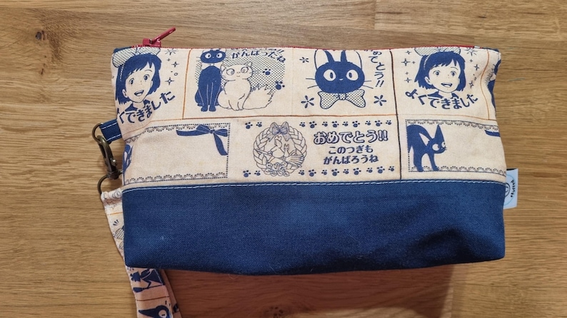 May include: A blue and cream fabric pouch with a zipper closure. The pouch features a repeating pattern of black cats, a white cat, and Japanese text. The text reads "できた!" and "おめでとう!!".