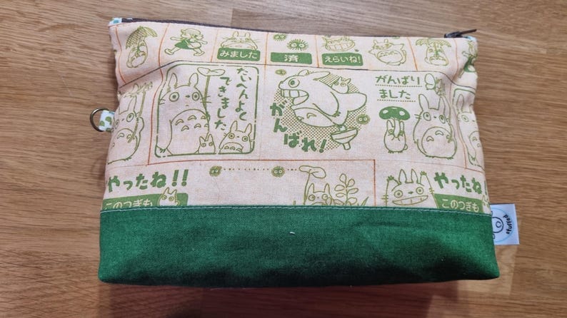 May include: A green and beige zippered pouch with a cartoon character print. The pouch features Japanese text and illustrations of a flying creature with large ears.