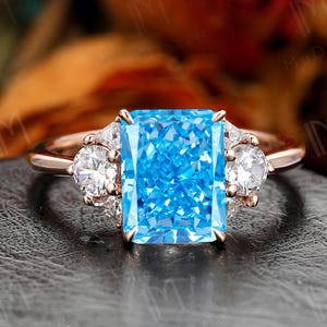 May include: Rose gold ring featuring a large, rectangular, bright blue gemstone as the centerpiece. Two smaller, round, clear gemstones flank the main stone. The ring is set against a dark background, with a blurred floral backdrop.