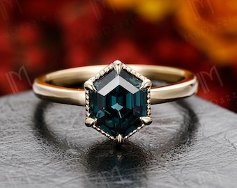 Könnte beinhalten: Ein goldener Ring mit einem sechseckigen, dunkelgrünen Edelstein, der in einer goldenen Fassung sitzt. Der Ring liegt auf einer grauen Oberfläche.
