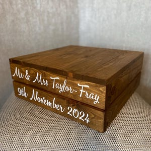 May include: Two-tiered wooden cake stand with the text "Mr. & Mrs. Taylor-Fray 9th November 2024" in white lettering.