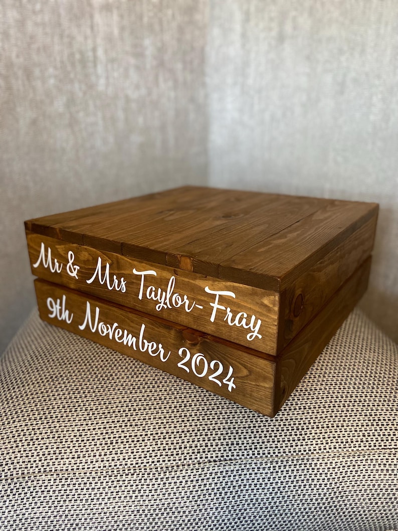 May include: Two-tiered wooden cake stand with the text "Mr. & Mrs. Taylor-Fray 9th November 2024" in white lettering.
