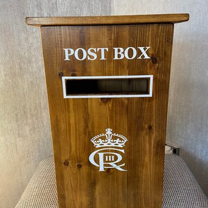 May include: A wooden post box with a white inscription that reads "POST BOX". The box has a white crown and the letters "C R" with a Roman numeral "III" below the letters.