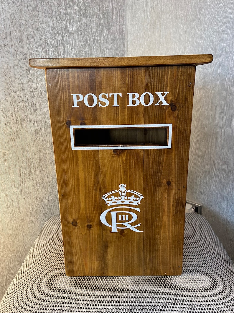 May include: A wooden post box with a white inscription that reads "POST BOX". The box has a white crown and the letters "C R" with a Roman numeral "III" below the letters.