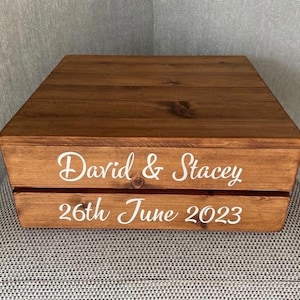 May include: A wooden box with a brown stain. The box has two levels and is decorated with white lettering that reads "David & Stacey 26th June 2023".