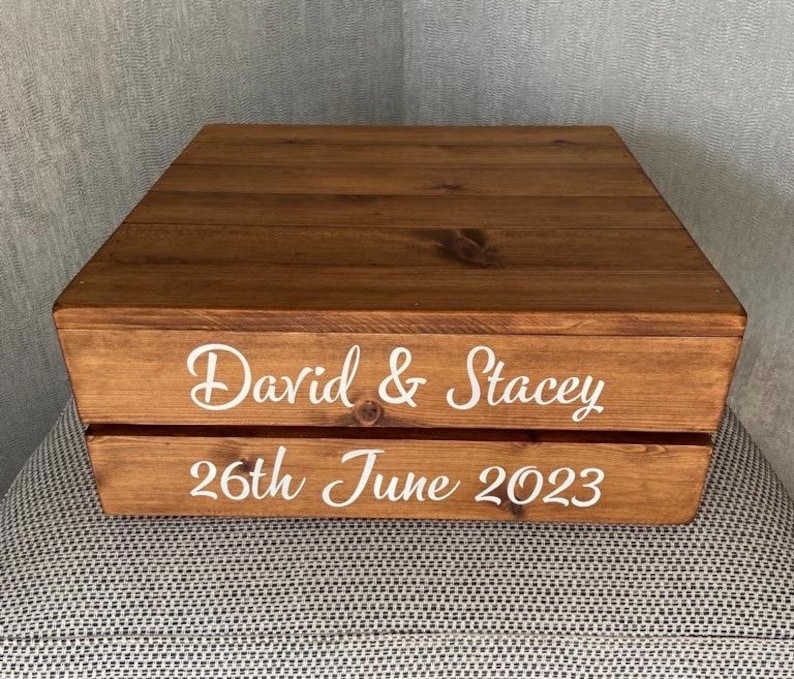 May include: A wooden box with a brown stain. The box has two levels and is decorated with white lettering that reads "David & Stacey 26th June 2023".