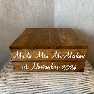 May include: A two-tiered wooden box with a brown stain. The top tier has a smooth surface and the bottom tier has the text "Mr & Mrs McMahon" and "1st November 2024" in white lettering.