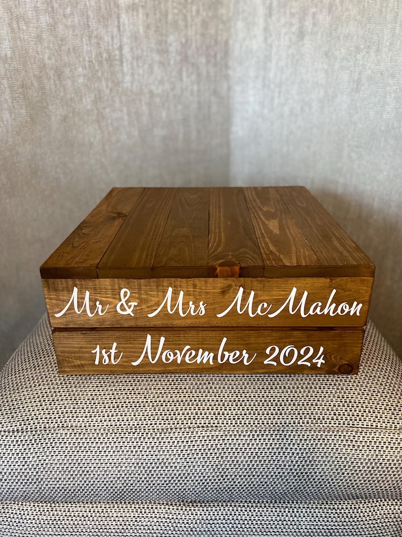 May include: A two-tiered wooden box with a brown stain. The top tier has a smooth surface and the bottom tier has the text "Mr & Mrs McMahon" and "1st November 2024" in white lettering.