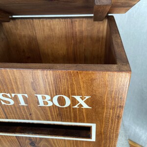 May include: A wooden post box with a hinged lid, a slot for mail, and the words "POST BOX" printed in white on the front. The box is stained a dark brown color and has a royal crest with a crown on the bottom.