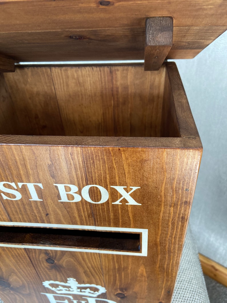 May include: A wooden post box with a hinged lid, a slot for mail, and the words "POST BOX" printed in white on the front. The box is stained a dark brown color and has a royal crest with a crown on the bottom.