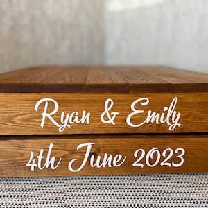 May include: A two-tiered wooden cake stand with the names "Ryan & Emily" and the date "4th June 2023" engraved in white.