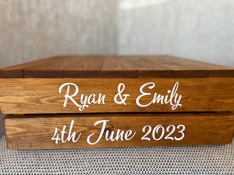 May include: A two-tiered wooden cake stand with the names "Ryan & Emily" and the date "4th June 2023" engraved in white.