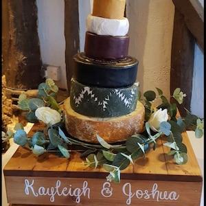 May include: A wooden crate wedding cake stand with the names "Kayleigh & Joshua" and the date "7th October 2022" written on it. The stand is decorated with greenery and a cheese wedding cake.