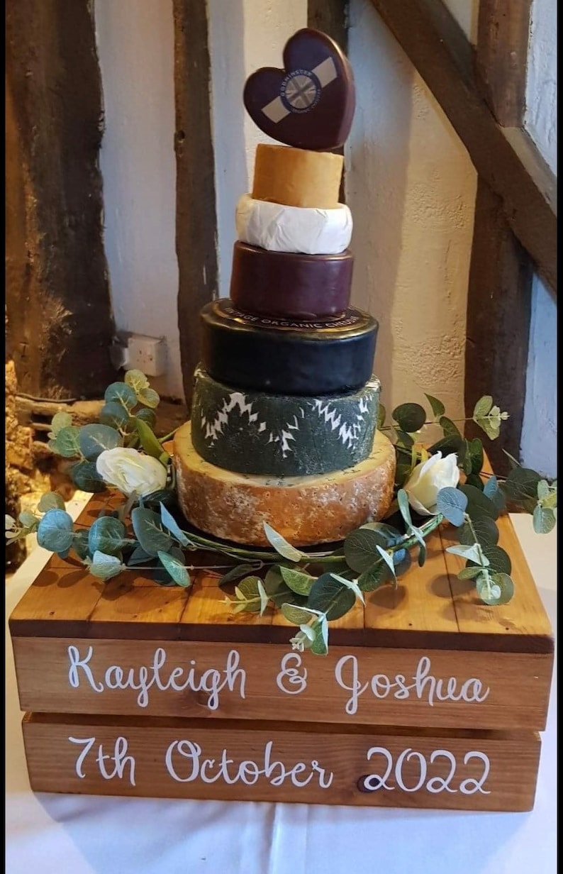 May include: A wooden crate wedding cake stand with the names "Kayleigh & Joshua" and the date "7th October 2022" written on it. The stand is decorated with greenery and a cheese wedding cake.