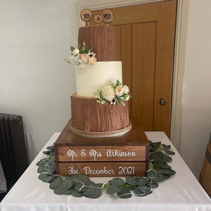 May include: A three-tiered wedding cake with a rustic woodsy theme. The cake is decorated with brown and white frosting and topped with wooden signs that say "Mr. & Mrs.". The cake is sitting on a wooden crate with the text "Mr. & Mrs. Atkinson 31st December 2021".