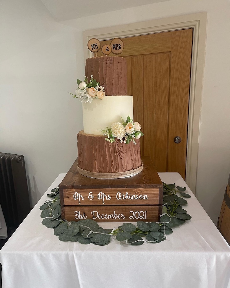 May include: A three-tiered wedding cake with a rustic woodsy theme. The cake is decorated with brown and white frosting and topped with wooden signs that say "Mr. & Mrs.". The cake is sitting on a wooden crate with the text "Mr. & Mrs. Atkinson 31st December 2021".