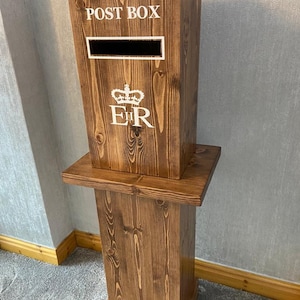May include: A wooden post box with a royal crest and the words "POST BOX" on the front. The post box is on a wooden stand.